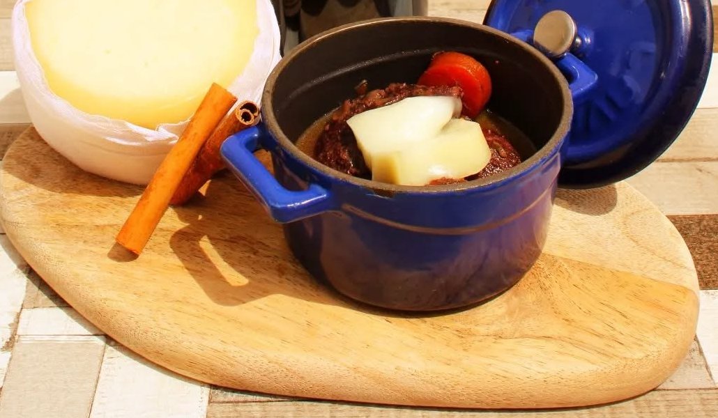 Caçarola azul com bochecha de porco preto, cenoura e lascas de queijo, ao lado de um queijo da serra e de um pau de canela, sobre uma tábua de madeira