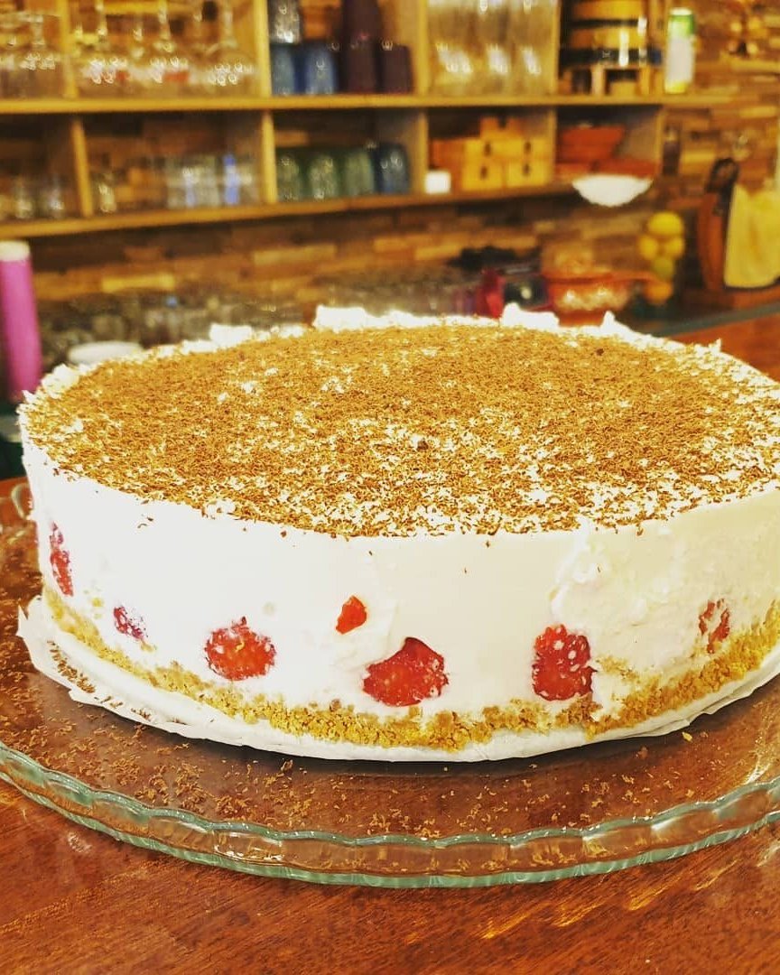 Bolo branco com morangos e raspas de chocolate sobre o balcão de madeira, com prateleiras de copos e loiça ao fundo